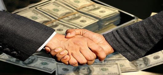 Handschlag mit vollem Geldkoffer im Hintergrund. Symbolbild für die Abhängigkeit der US-Abgeordneten vom Geld